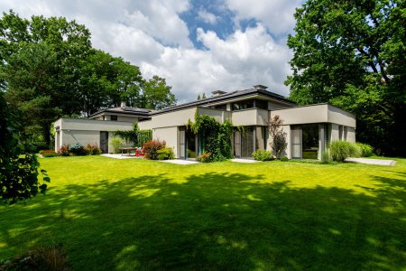 Nowoczesna rezydencja wybudowana przez generalnego wykonawcę New-House. Widoczna jasna elewacja, duże przeszklenia oraz w pełni zagospodarowany ogród z trawnikiem i nasadzeniami